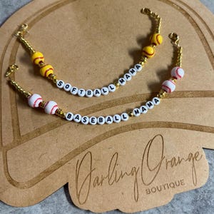 May include: Two gold-colored beaded bracelets with white letter beads spelling "SOFTBALL MAMA" and "BASEBALL MAMA". The bracelets feature baseball and softball-themed beads. The background is a brown surface with the text "Darling Orange Boutique".