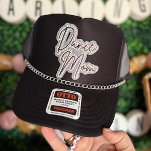 Peut inclure: Casquette de camionneur noire avec une chaîne argentée et un patch en strass blanc qui dit "Dance Mom". La casquette a un dos en maille noire et un devant en mousse noire.