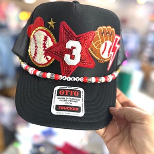 May include: Black trucker cap featuring baseball-themed appliqués: a red star with the number 3, a baseball, and a baseball glove. The cap includes a beaded band with the name "ALEXO". Made from foam fabric, it has a snap closure.