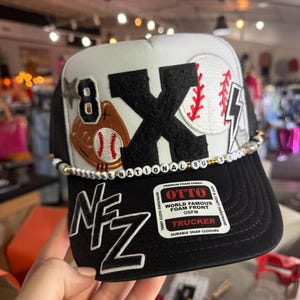May include: Black and white trucker hat with a baseball theme. The hat features a large black "X" and baseball graphics. The brim is black, and the crown is white with a black mesh back. The text "8" and "NFZ" are also visible.