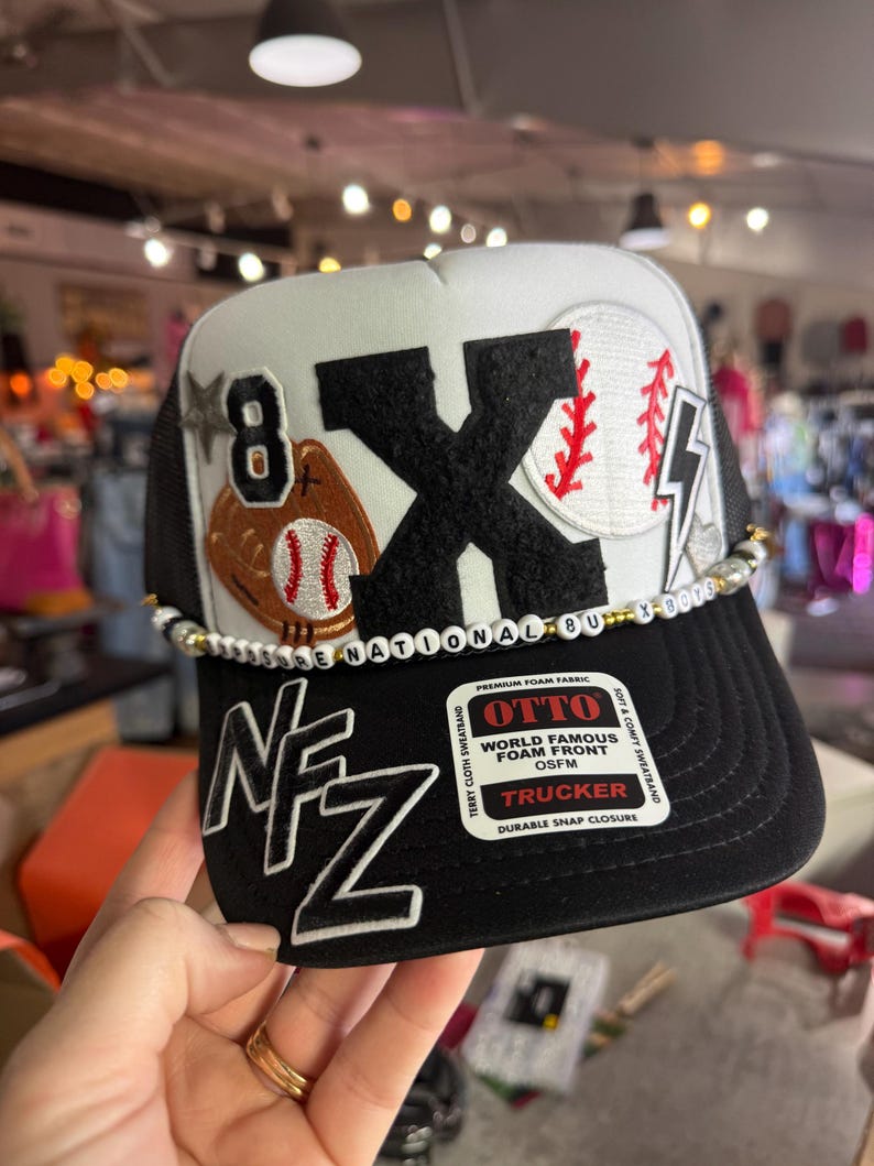May include: Black and white trucker hat with a baseball theme. The hat features a large black "X" and baseball graphics. The brim is black, and the crown is white with a black mesh back. The text "8" and "NFZ" are also visible.
