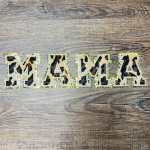 May include: Four fabric letters spell out the word "MAMA". The letters have a leopard print pattern with a gold glitter border and a white outline. The letters are arranged horizontally on a wooden surface.