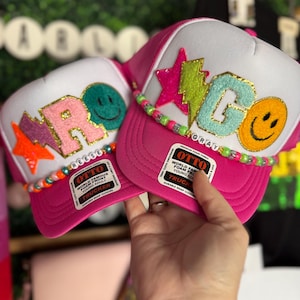 May include: Two pink and white trucker hats with colorful letters spelling out "REESE" and "GRAY". Each hat has a smiley face patch, a star patch, and a beaded trim.