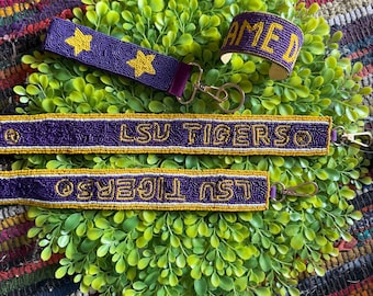 May include: Purple beaded accessories, including a keychain with gold stars, a bracelet with the word "AMED", and two keychains with "LSU TIGERSO" in gold lettering. The items are displayed on a bed of green foliage.