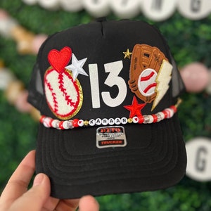 May include: Black trucker hat with a white number 13, a red heart, a baseball, a baseball glove, and a lightning bolt. The hat has a white beaded band with the name "Kanaan" spelled out in white letters. The hat also has a red and white "Otto" label with the text "World Famous Foam Front Trucker".