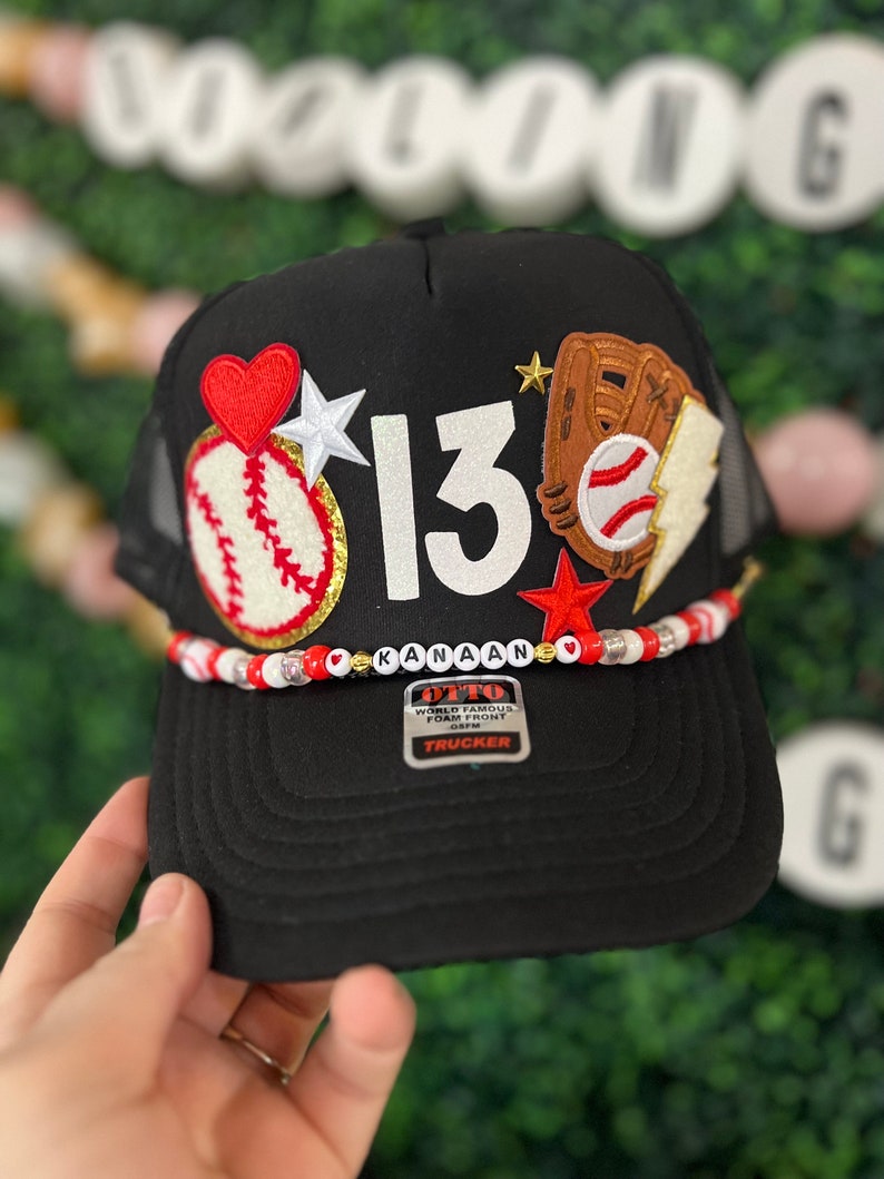 May include: Black baseball cap with a mesh back, decorated with a red heart, white star, baseball, glove, lightning bolt, and the number 13. The name "Kanaan" is spelled out on the brim with beads. The cap has a "Trucker" tag.