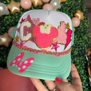 May include: A mint green and white trucker hat with a pink and gold chain. The hat features a pink Minnie Mouse design, a pink lightning bolt, and a letter "C". The brim has a pink bow.