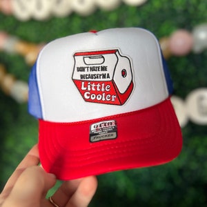 May include: A white, blue and red trucker hat with a patch that says "Don't hate me because I'm a Little Cooler". The hat has a mesh back and a foam front.