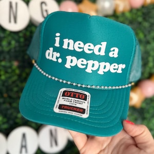 May include: Teal trucker hat with a white beaded trim and a white graphic that says "i need a dr. pepper".
