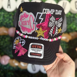 May include: Black trucker hat with a pink and white beaded trim. The hat has several patches including a pink megaphone with the word "RORY" on it, a pink star with a black and white checkered bow, a gold star, and a lightning bolt. The hat also has a white patch with the text "CHEER MAMA" and a black patch with the text "COMP DAY". The hat has a black label with the text "OTTO WORLD FAMOUS FOAM FRONT OSFM TRUCKER DURABLE SNAP CLOSURE".