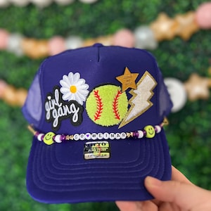 May include: Purple trucker hat with a white daisy, a yellow softball, a gold star with the text "Doing My Best", and a gold lightning bolt. The hat has a purple and white beaded band with smiley faces and the text "Go Weimar".