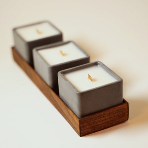 May include: Three gray ceramic candle holders with white candles and wooden wicks, arranged on a wooden tray.
