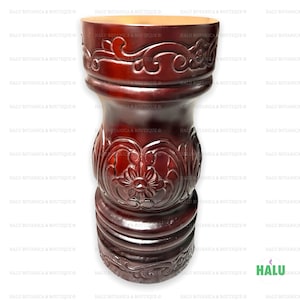May include: A dark brown wooden pedestal with intricate carvings. The pedestal has a round top and a cylindrical base. The carvings are in a floral pattern.