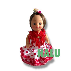 May include: A doll in a red fringed shawl and a white dress with red polka dots. The doll has brown hair and a floral hairpiece. The word "HALU" is printed in green on the dress.