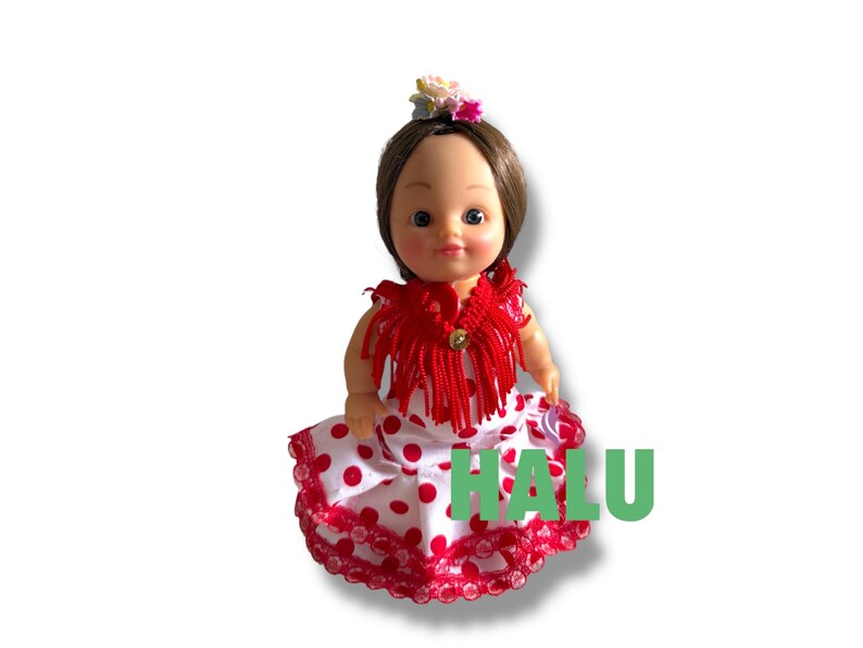 May include: A doll in a red fringed shawl and a white dress with red polka dots. The doll has brown hair and a floral hairpiece. The word "HALU" is printed in green on the dress.