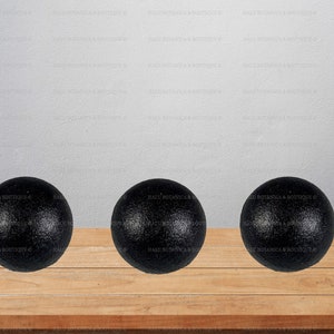 Puede incluir: Tres bolas de masaje negras con superficie texturizada dispuestas sobre una superficie de madera. Las bolas esféricas parecen estar hechas de un material denso. El fondo es una pared gris claro.