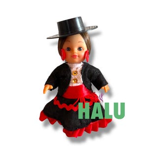 May include: A doll in a traditional Spanish flamenco costume. The doll is wearing a black hat, a black jacket over a white blouse, a red sash, and a black skirt with red trim. The doll has red earrings and the word "HALU" in green.