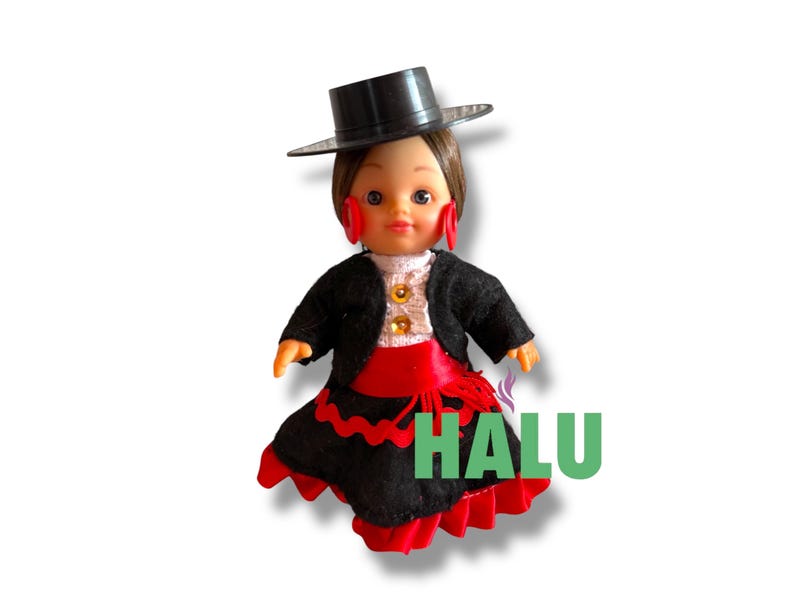 May include: A doll in a traditional Spanish flamenco costume. The doll is wearing a black hat, a black jacket over a white blouse, a red sash, and a black skirt with red trim. The doll has red earrings and the word "HALU" in green.