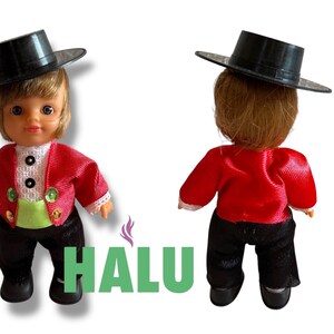 May include: Two small dolls in traditional dress. Each doll wears a black hat, red jacket, black trousers, and black shoes. One doll faces forward, and the other faces away. The dolls are on a white background.
