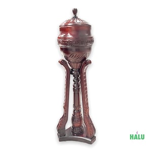 May include: A dark reddish-brown wooden urn with a lid, set on a tall, ornate stand. The urn has carved floral details around the middle and on the stand's legs. The stand is approximately 40 inches tall.