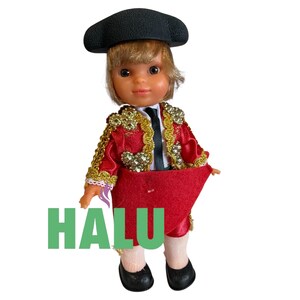 May include: A doll dressed as a matador, wearing a black hat, a red and gold jacket, and a red cape. The doll has blonde hair and is wearing black shoes. The word "HALU" is printed in green.