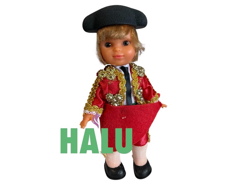 May include: A doll dressed as a matador, wearing a black hat, a red and gold jacket, and a red cape. The doll has blonde hair and is wearing black shoes. The word "HALU" is printed in green.