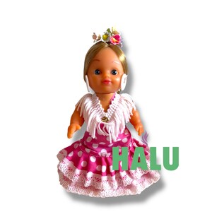 May include: A doll in a traditional Spanish flamenco dress. The doll has blonde hair, a floral headpiece, and white earrings. The dress is pink with white polka dots and lace trim. The word "HALU" is printed in green.