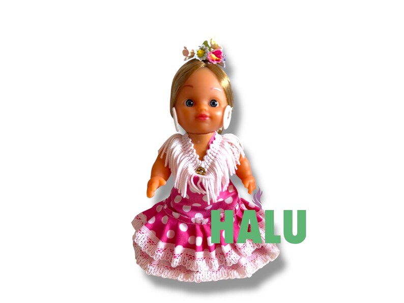 May include: A doll in a traditional Spanish flamenco dress. The doll has blonde hair, a floral headpiece, and white earrings. The dress is pink with white polka dots and lace trim. The word "HALU" is printed in green.