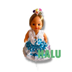 May include: A doll in a traditional Spanish flamenco dress. The doll has blonde hair and a floral headpiece. The dress is blue with white polka dots and lace trim. The word "HALU" is printed in green.