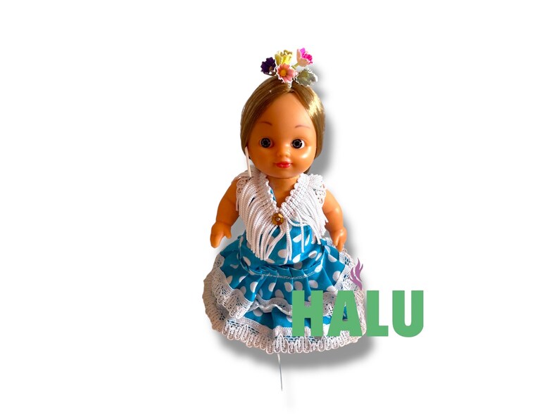 May include: A doll in a traditional Spanish flamenco dress. The doll has blonde hair and a floral headpiece. The dress is blue with white polka dots and lace trim. The word "HALU" is printed in green.