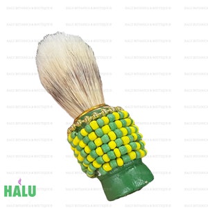 Puede incluir: Un cepillo de afeitar con cerdas blancas y marrones con un mango de madera verde y amarillo. El mango está adornado con cuentas amarillas y verdes y una banda dorada alrededor de la base de las cerdas.