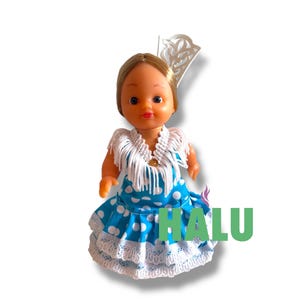 May include: A doll in a blue polka-dot dress with white lace trim and a white fringed shawl. The doll has blonde hair and a decorative headpiece. The word "HALU" is written in green letters.