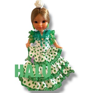 May include: A doll wearing a traditional flamenco dress with white fabric and green polka dots. The dress has ruffled layers and green trim. The doll has brown hair, green earrings, and a white flower in her hair. The word "HALU" is printed in green.