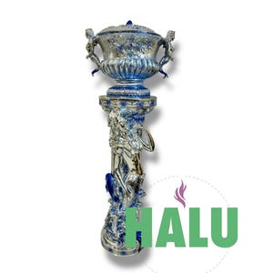 May include: A silver and blue decorative urn with a lid, supported by a detailed pedestal. The urn features handles and floral designs. The pedestal is adorned with sculpted figures and blue accents. The word "HALU" is visible in green.