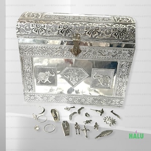 Op de afbeelding: Een zilveren metalen schatkist met ingewikkelde bloem- en olifantmotieven. De kist is open en bevat een verscheidenheid aan kleine zilveren metalen charmes, waaronder een kruis, een sleutel, een halve maan, een ster, een blad en een kist.