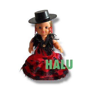 May include: A doll dressed in a red dress with black polka dots and lace trim, a black shawl, and a black hat. The doll has blonde hair and red earrings. The word "HALU" is printed in green.