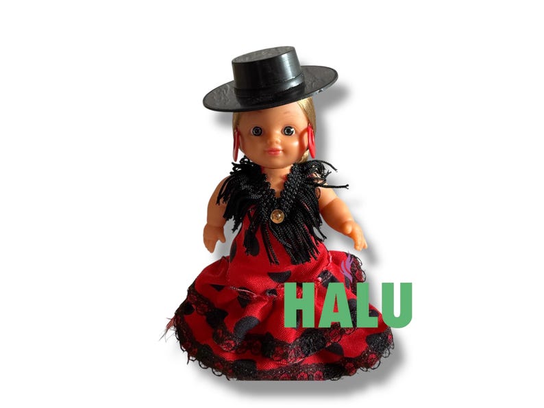 May include: A doll dressed in a red dress with black polka dots and lace trim, a black shawl, and a black hat. The doll has blonde hair and red earrings. The word "HALU" is printed in green.