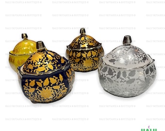 Pode incluir: Quatro taças de cerâmica com tampa, bordas recortadas e padrões florais. Uma é amarela com ouro, uma azul-escura com ouro, uma preta com ouro e uma branca com prata. Cada taça possui uma tampa a condizer com um pequeno puxador.