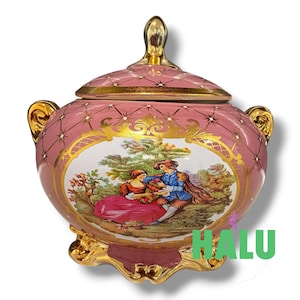 May include: A pink porcelain lidded jar featuring a gold lattice pattern with star accents. The center displays a colorful scene of two figures in a landscape. The jar has gold handles, a gold finial on the lid, and a gold base with ornate feet.