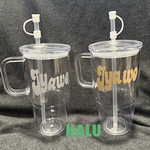 May include: Two clear plastic tumblers with handles, lids, and straws. Each tumbler has the word "Jyawo" in a different color, one silver and one gold. The tumblers are on a grey surface.