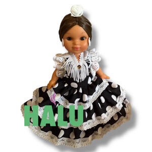 May include: A doll wearing a black and white polka dot dress with white lace trim. The doll has brown hair, a white flower hair accessory, and white earrings. The word "HALU" is written in green letters on the dress.
