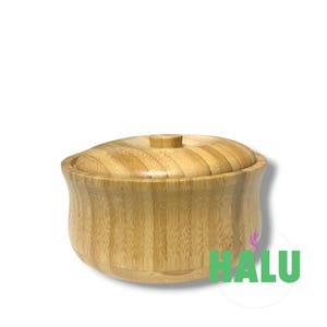 Puede incluir: Un recipiente de bambú de color marrón claro con tapa a juego, que presenta una forma redondeada y estriada con una textura de veta de madera visible. La palabra HALU está impresa en letras verdes en la parte inferior derecha, junto a un icono de llama morada.