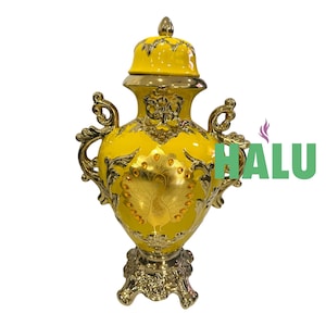 May include: A yellow decorative vase with a lid, adorned with gold accents and a peacock design. The vase has ornate handles and a detailed base. The word "HALU" is visible in green.