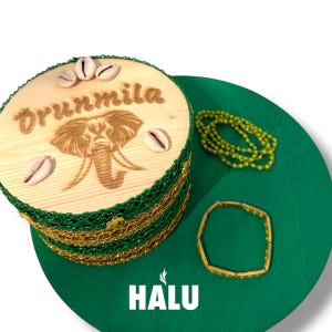 Könnte beinhalten: Eine runde Holzbox mit einer Elefanten-Schnitzerei und dem Namen "Oruumila". Die Box ist mit Muscheln und grünen und goldenen Verzierungen verziert. Eine grüne Perlenkette und ein Armband sind ebenfalls vorhanden. Das Wort "HALU" steht unten.