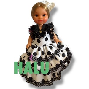 May include: A doll wearing a traditional Spanish flamenco dress. The dress is white with large black polka dots and black lace trim. The doll has blonde hair and a decorative headpiece. The word "HALU" is printed in green on the dress.
