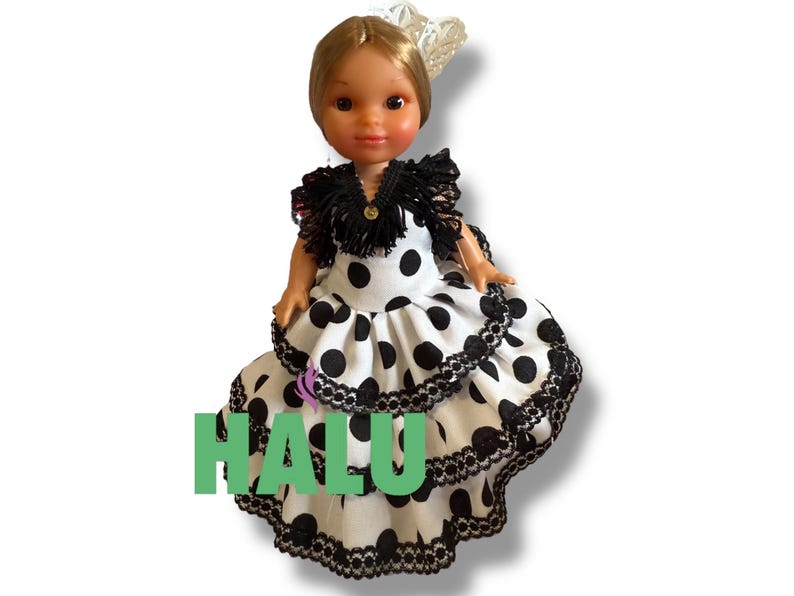 May include: A doll wearing a traditional Spanish flamenco dress. The dress is white with large black polka dots and black lace trim. The doll has blonde hair and a decorative headpiece. The word "HALU" is printed in green on the dress.
