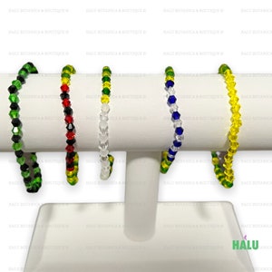 May include: Five beaded bracelets in different colours are displayed on a white stand. The bracelets feature green, red, clear, blue, and yellow beads. The bracelets are made of small, faceted beads strung together.