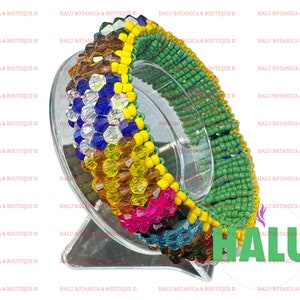 Peut inclure: Un bracelet en perles colorées avec des perles vertes, jaunes, bleues et brunes. Le bracelet est sur un présentoir transparent. Le mot "HALU" est visible dans le coin inférieur droit de l'image.