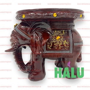 May include: A dark brown elephant-shaped side table with a round top. The table features intricate carvings, gold accents, and a decorative saddle. The elephant's head is detailed with tusks and a jeweled headpiece. The text "HALU" is visible.