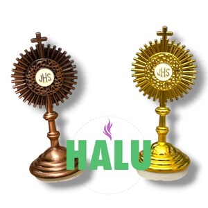 Puede incluir: Dos réplicas de custodia, una con acabado en bronce y otra en dorado, cada una con un disco central blanco con las letras JHS, un diseño de rayos y una pequeña cruz en la parte superior, montadas sobre una base circular.
